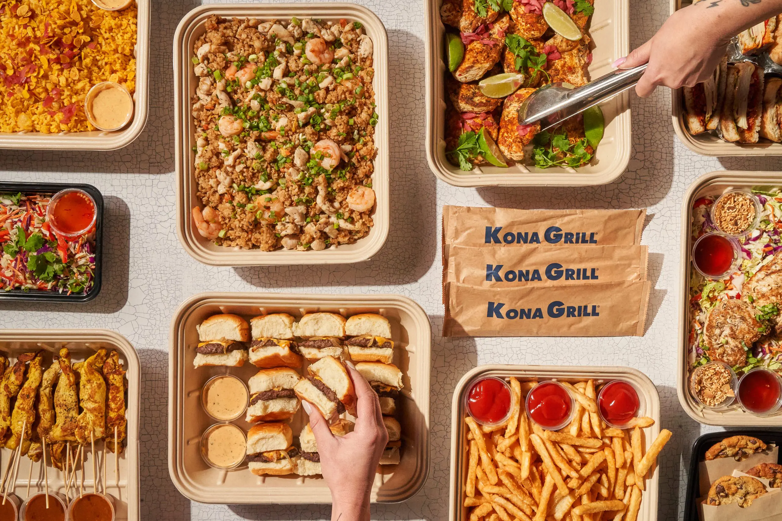 Overview of catering trays including chicken satay skewers, fried rice with shrimp and chicken, KG sliders with dipping sauce, French fries with ketchup, Asian slaw, and cookies. Two hands reach in-one taking a slider, another serving grilled chicken with tongs.