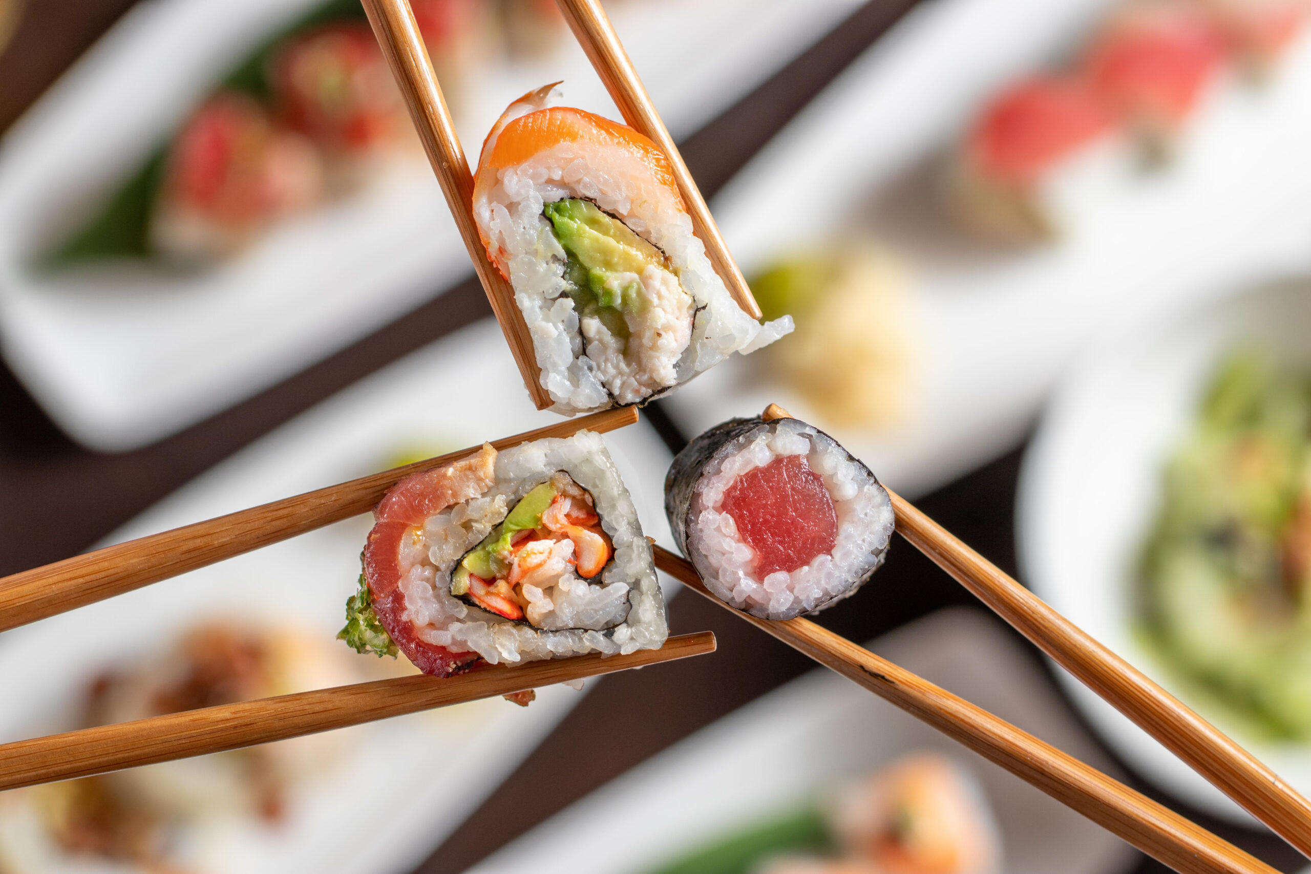 Three pieces of sushi held by chopsticks: a Rainbow Roll with salmon, avocado, crab salad; a Bama Roll with crab salad, cream cheese, jalapeño, tuna, and avocado; and a traditional Tuna Roll, viewed from above