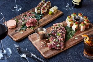 Two cutting boards of steak and sushi with a cocktail and a glass of red wine