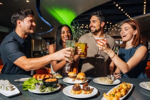 4 people raising their glasses in cheers over a selection of Kona Grill meals.