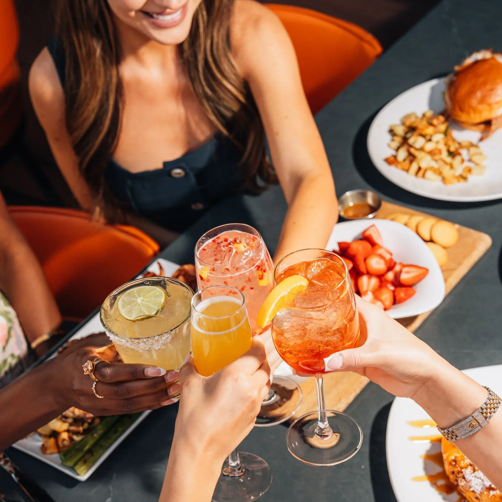 A group of friends gathered around a table at Kona Grill raise their glasses of for a toast. Each person holds in their hand a different style of beverage, and there is a spread of various entrees on the table.
