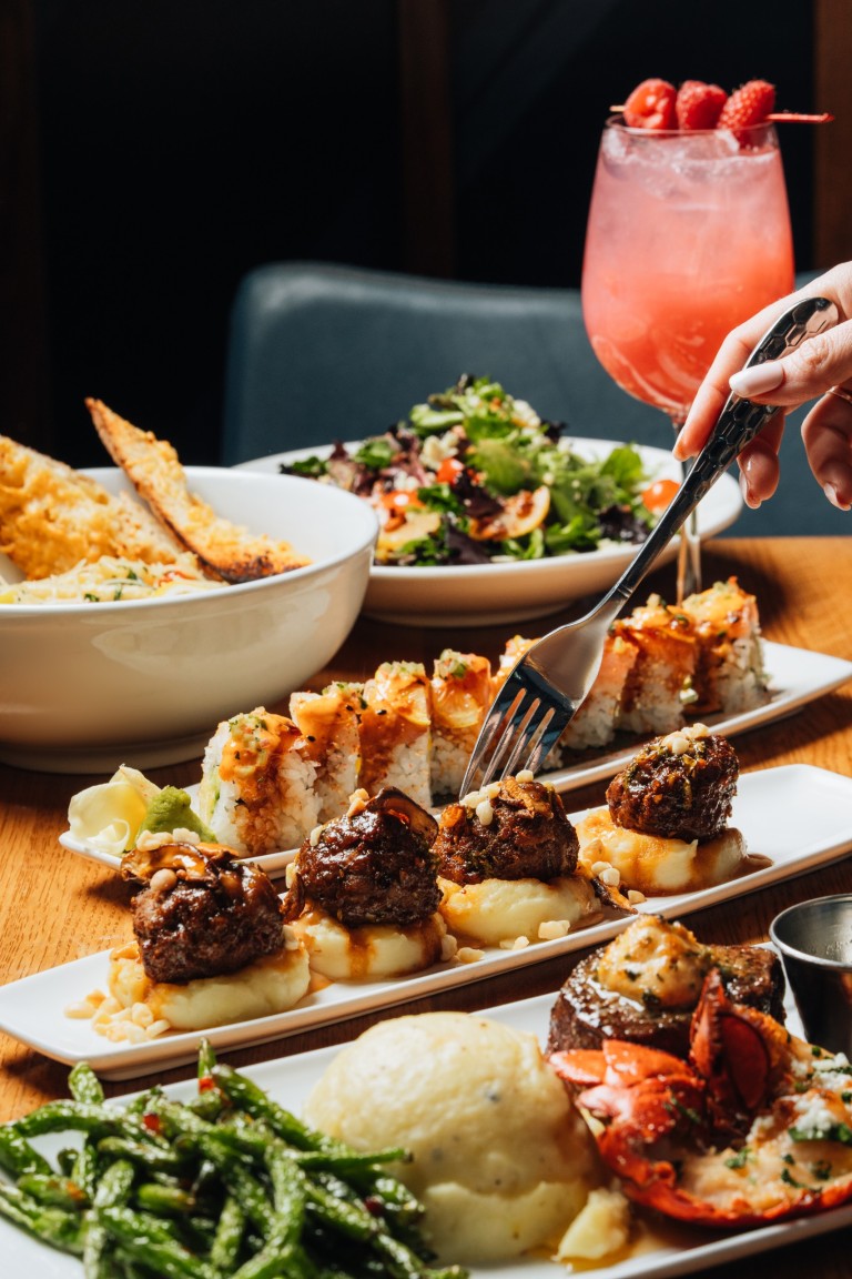 A variety of Kona Grill dishes and drinks set on a table, including sushi rolls, mini meatball skewers on rice, a lobster tail with mashed potatoes and green beans, mixed green salads, a pasta bowl, and a pink gin or cocktail garnished with berries. A person is using a knife and fork to serve from one of the plates.
